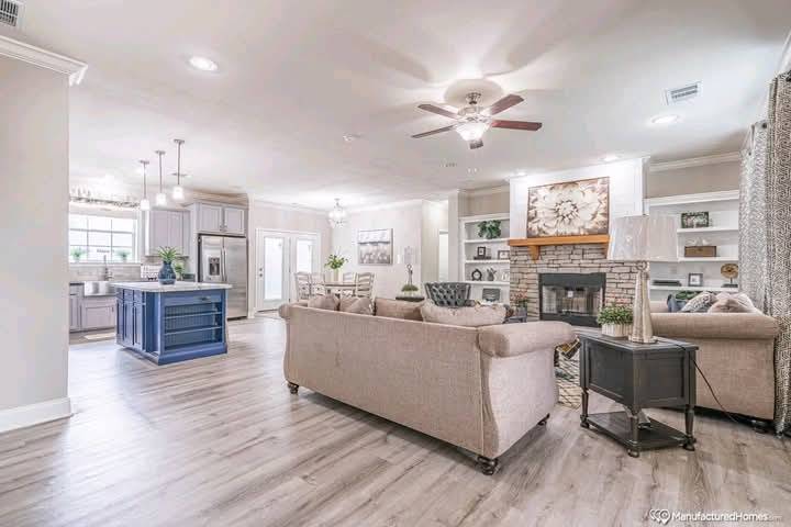 Spacious living area with beige sofas, a stone fireplace, and a ceiling fan. Open kitchen with a blue island and modern appliances. Bright, cozy atmosphere.