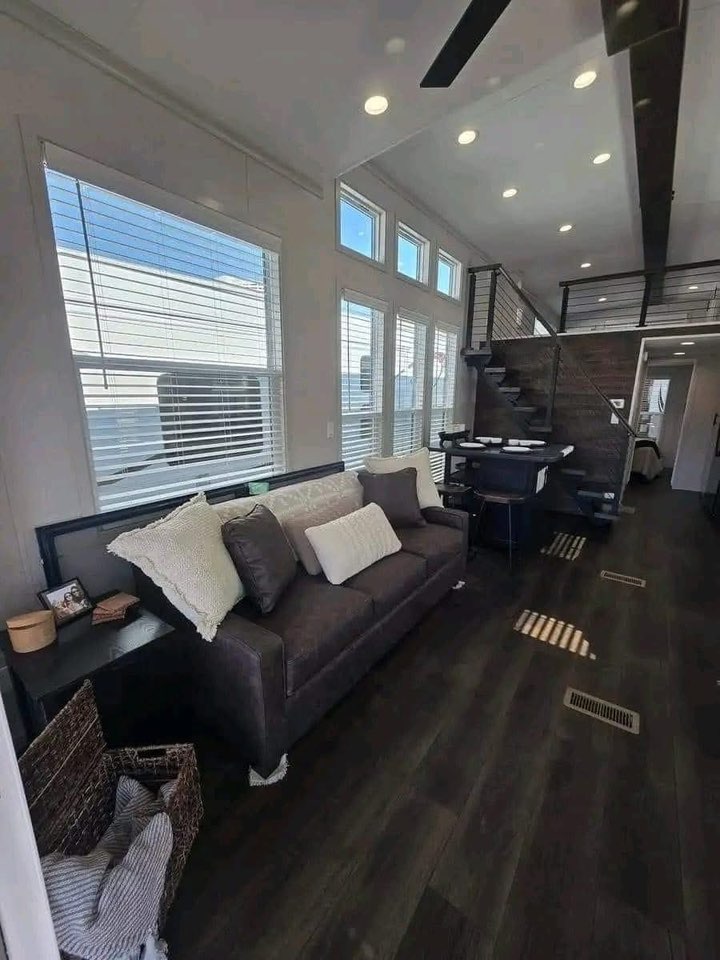 Modern tiny home interior with dark flooring, a cozy brown sofa with white cushions, and a staircase leading to a loft. Bright, airy atmosphere.