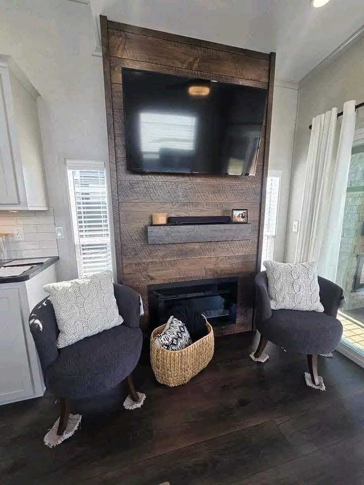 Cozy living room with two gray armchairs, white pillows, a large TV on a wooden accent wall, a small fireplace, and a wicker basket in between. Bright ambiance.