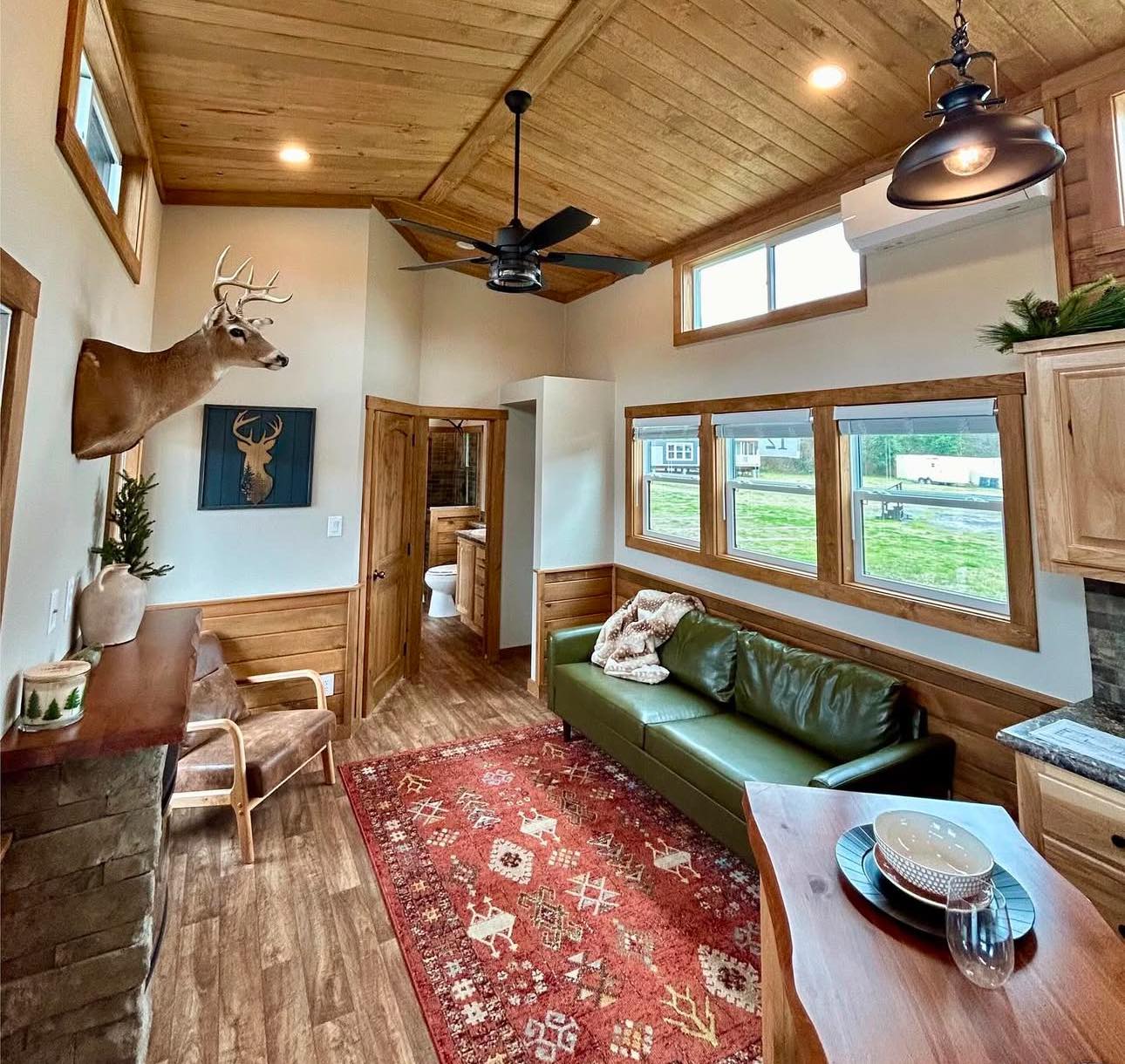 Cozy cabin interior with wood paneling, a mounted deer head, green leather sofa, and red patterned rug. Large windows and rustic decor create a warm ambiance.