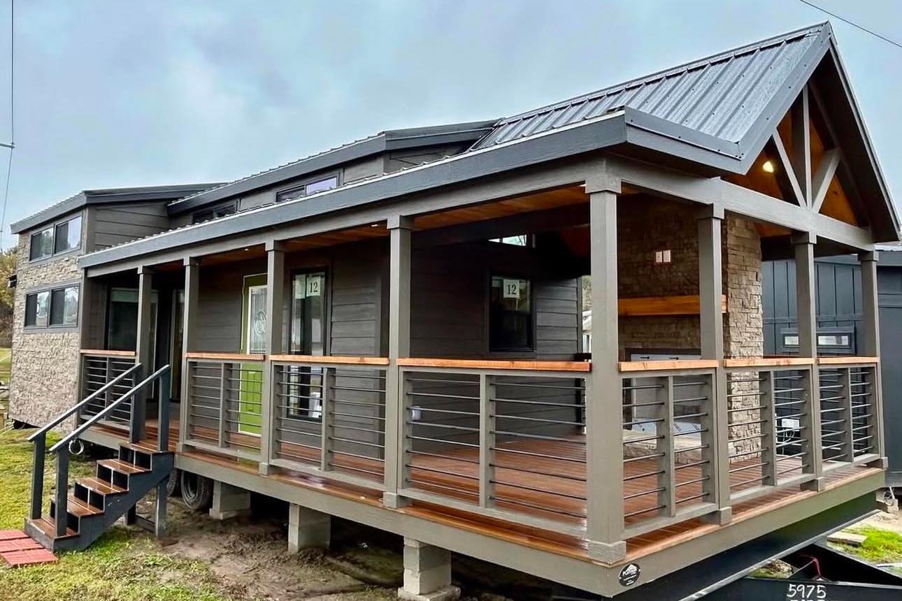 A modern tiny house with a metal roof and a stone accent wall. It features a covered porch with horizontal railings and wooden steps, set against a cloudy sky.