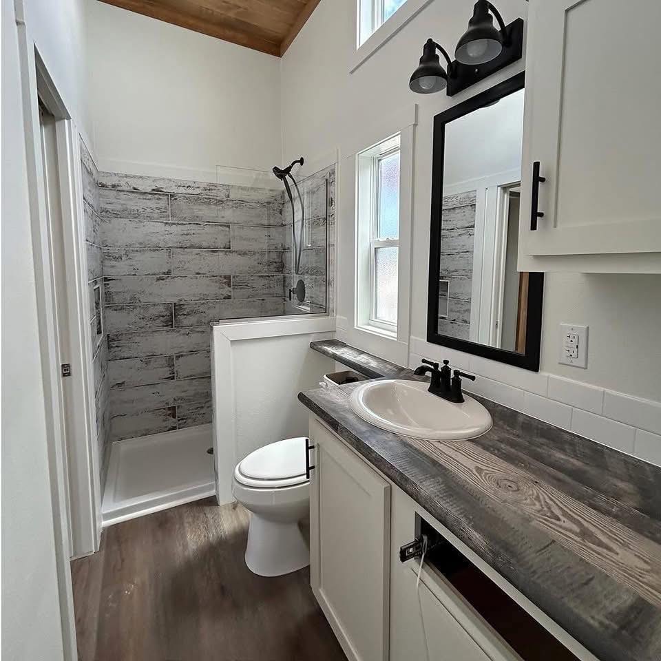 Compact bathroom with rustic wood accents. Features a shower with weathered tiles, a white vanity with a dark wood countertop, and a black-framed mirror.