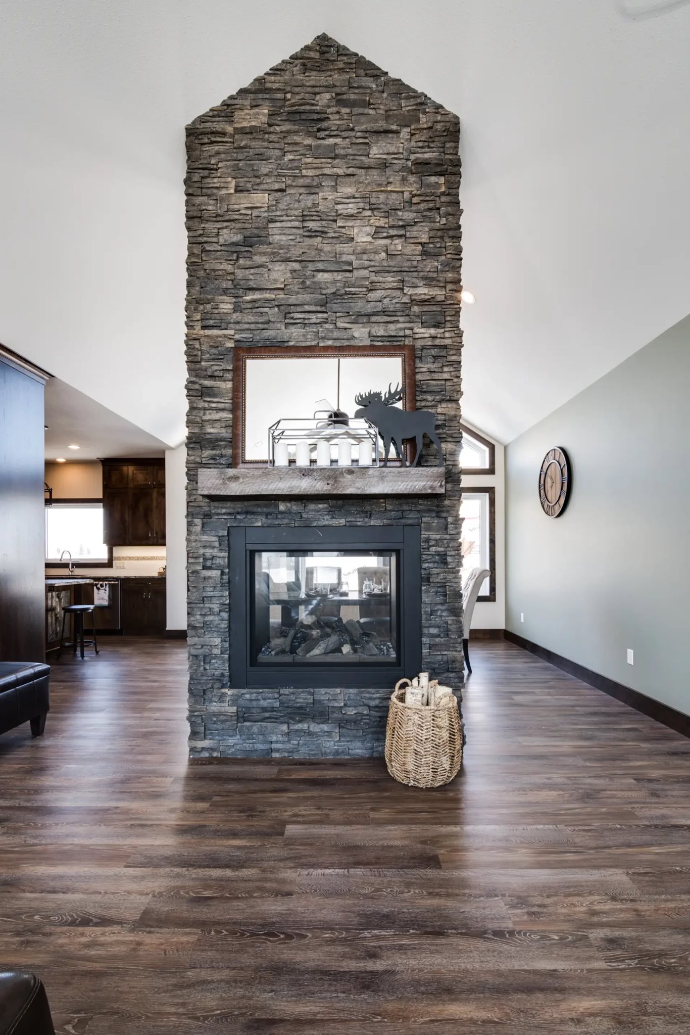 Modern living room with a tall stone fireplace, wooden floors, and soft lighting. A woven basket and decorative items add a cozy, rustic touch.