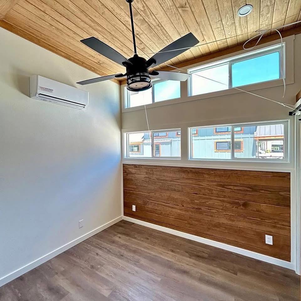 A minimalist room with wood panel accent wall, large windows, and neutral colors. Ceiling fan and air conditioner give a modern, cozy feel.
