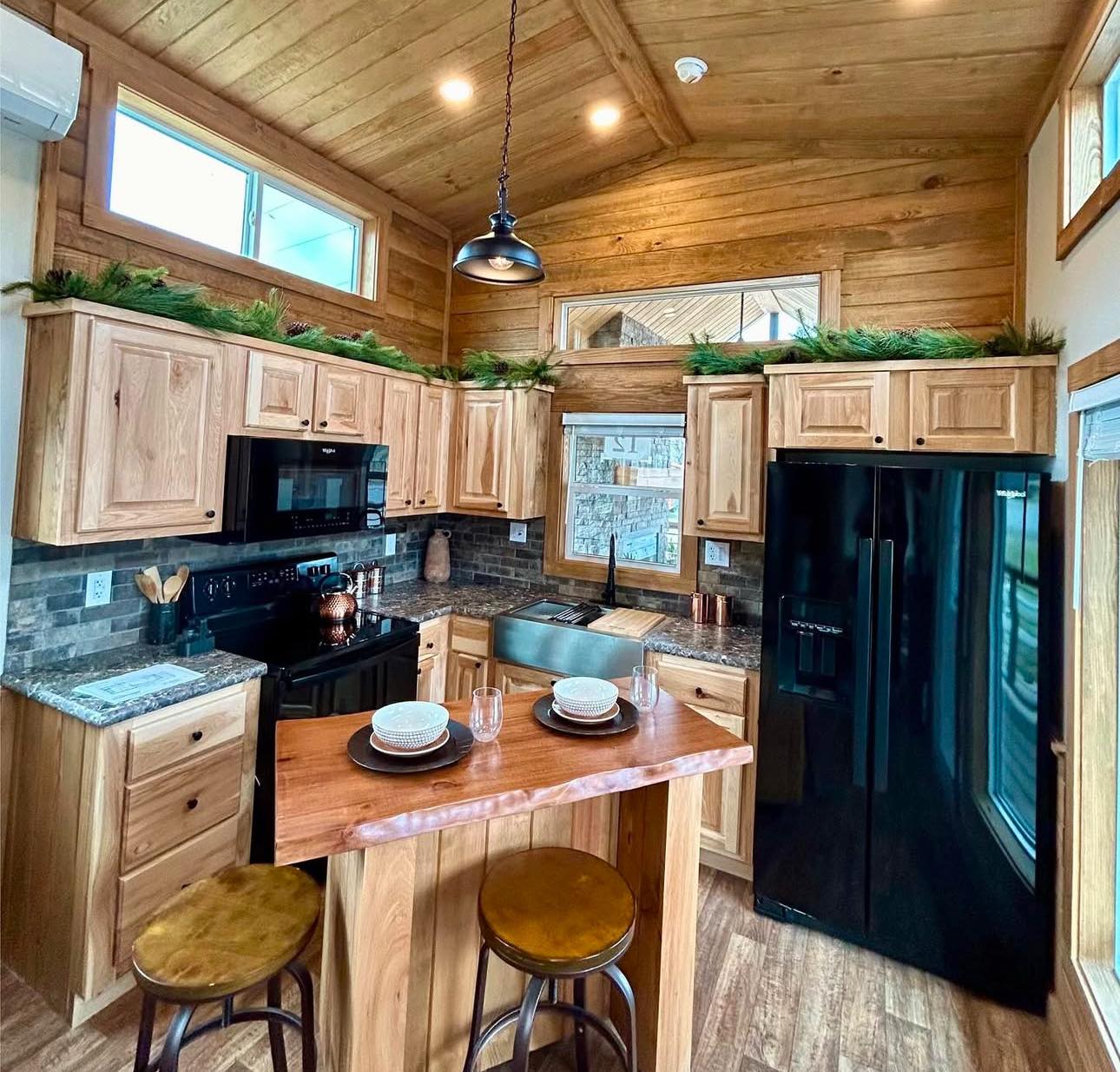 Cozy kitchen with wooden cabinets and ceiling, black appliances, and a granite countertop. A small wooden island has two stools and set plates, under a warm light.