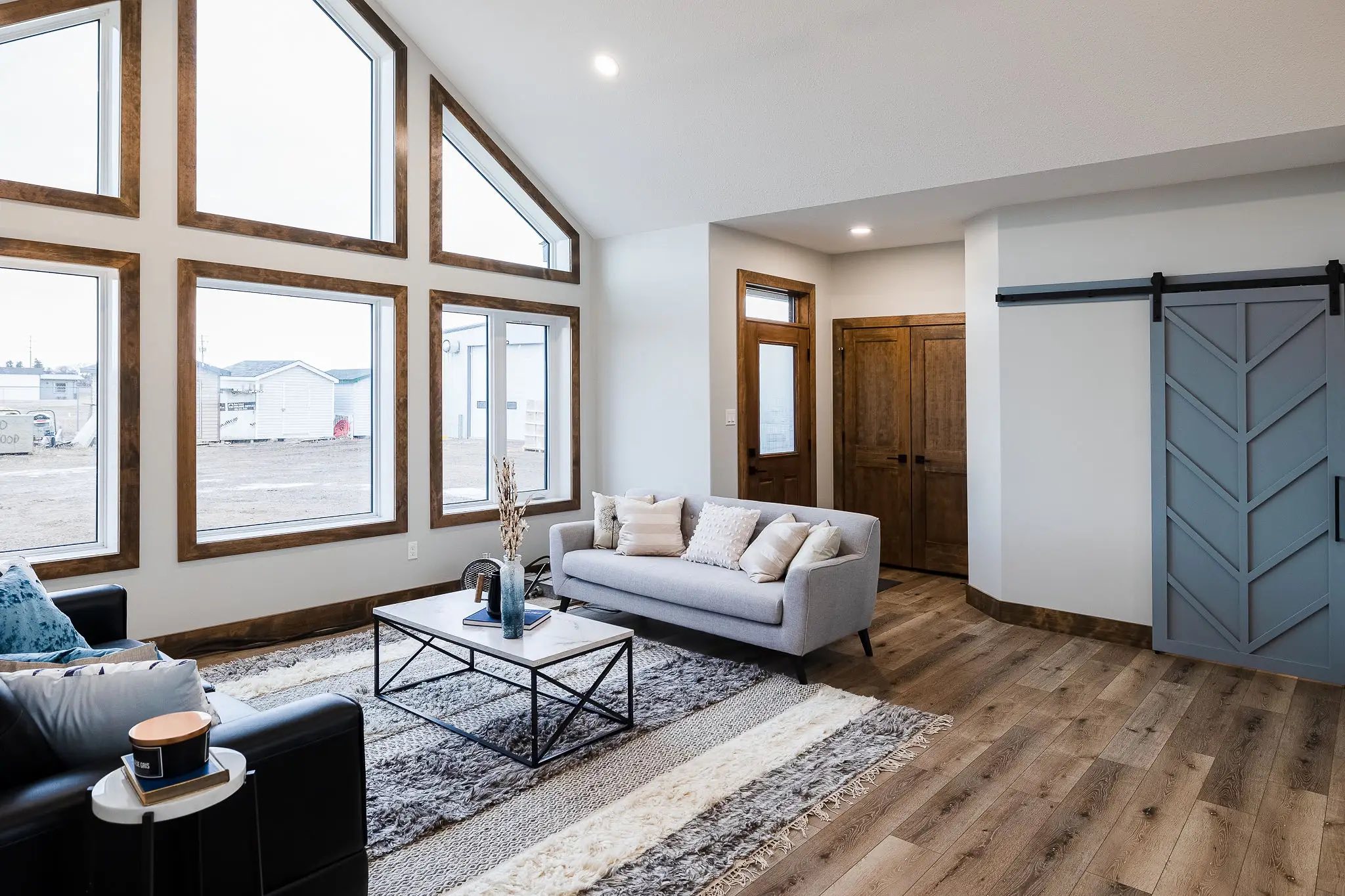 Modern living room with large triangular windows, a gray sofa, and a black armchair. Features a rustic sliding barn door and wooden floor, exuding warmth.