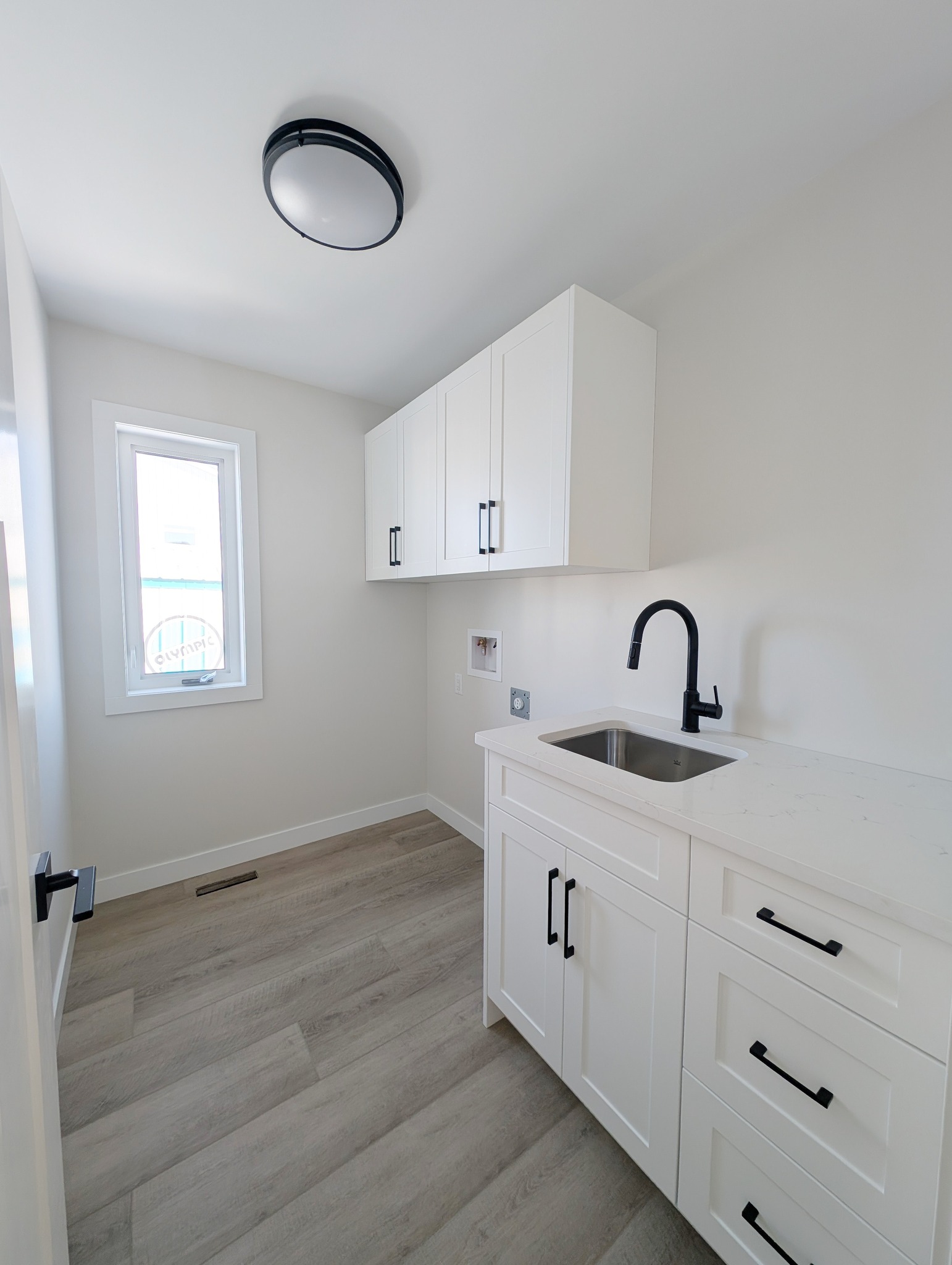 A minimalist laundry room with light wood flooring, white cabinets, and a black faucet over a sink. A window brings bright, natural light in.