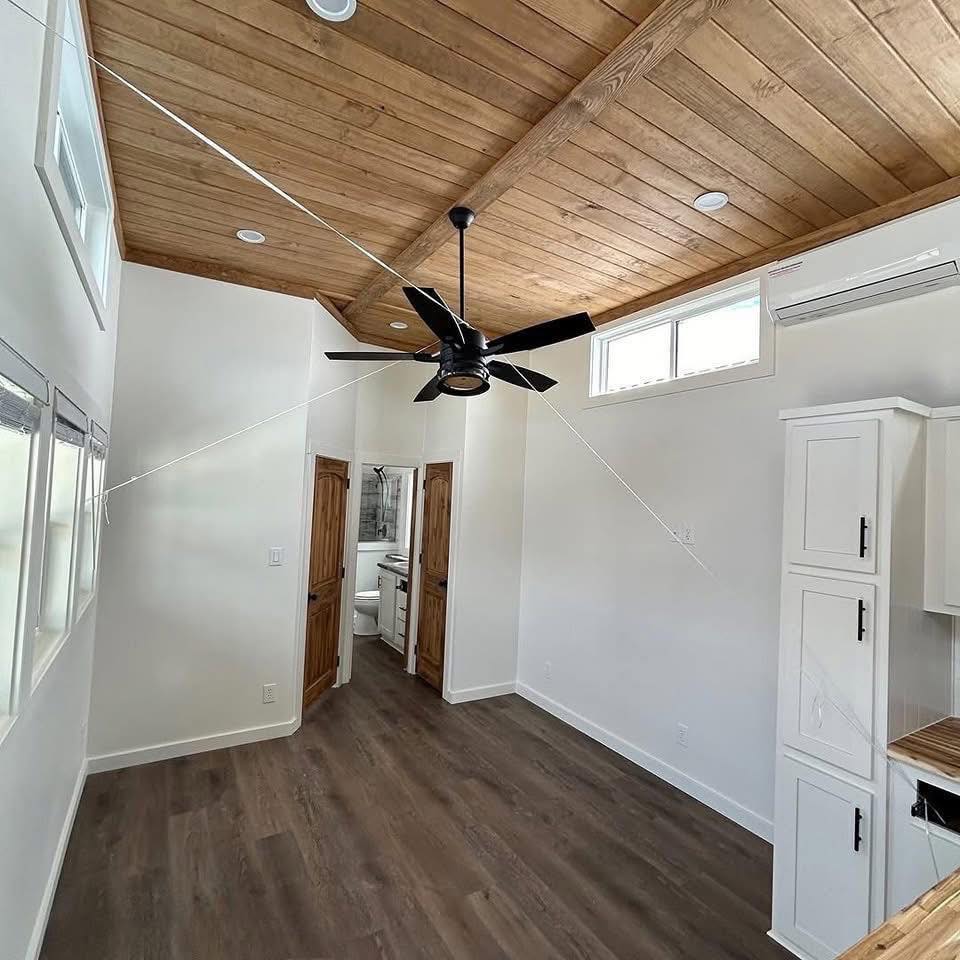 A small, bright room with a wooden ceiling and floor. There's a modern black ceiling fan, white walls, cabinets, and light filtering through high windows. The space feels airy and minimalist, leading to a small bathroom.
