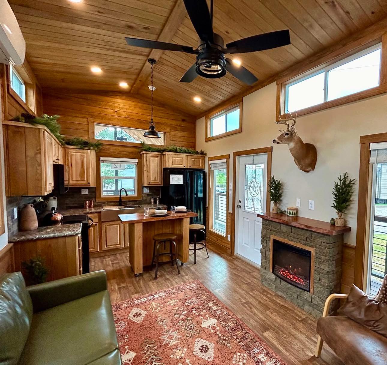 Cozy cabin interior with wooden ceiling and floors. Features a modern kitchen, a ceiling fan, a fireplace, and decorative mounted deer head. Warm, inviting atmosphere.