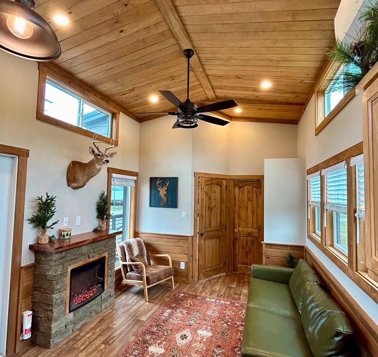 Cozy room with wood ceiling, stone fireplace, and a green sofa. Deer head mounted above the fireplace, warm lighting, rustic decor, and natural tones.