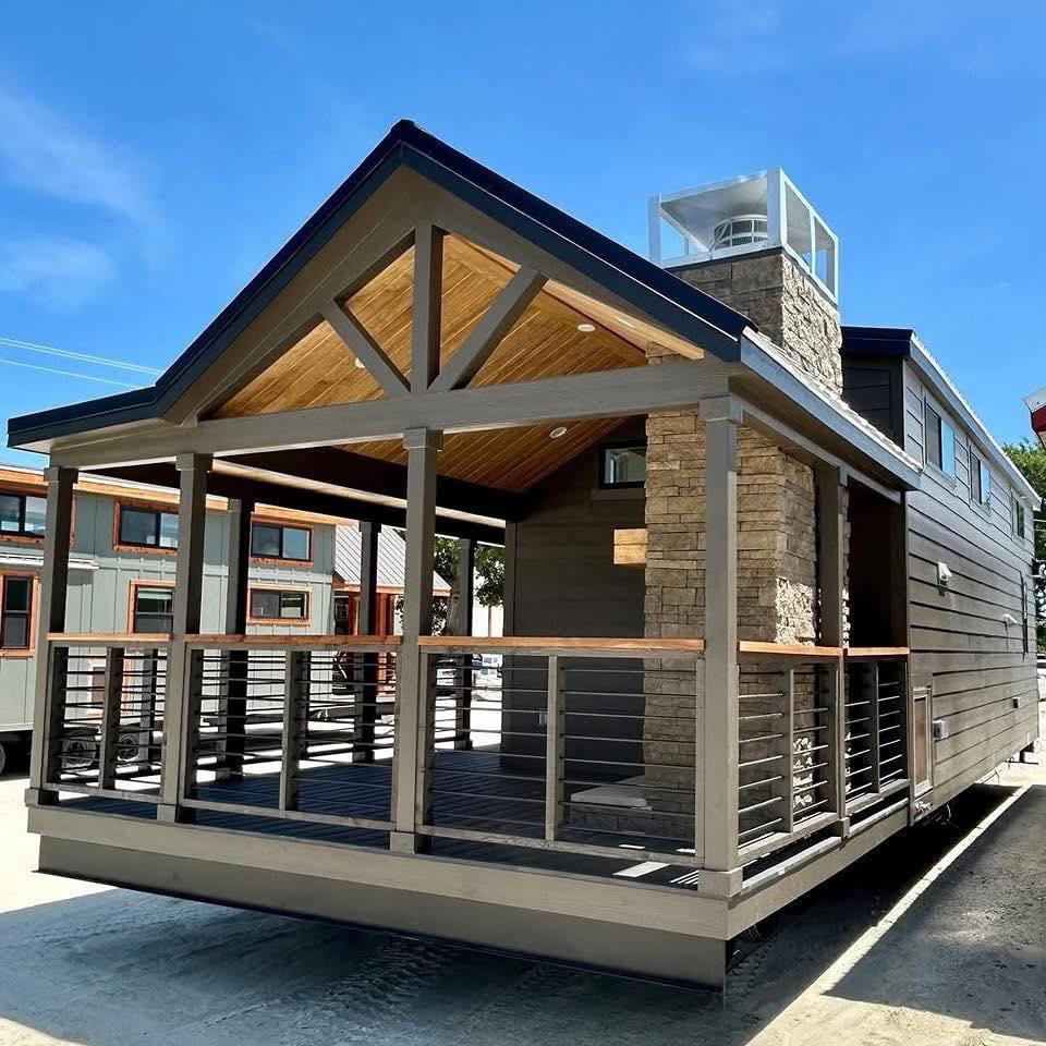 Modern tiny house with a front porch and stone chimney, featuring a gabled roof and wooden railings. Set in a sunny area with a clear blue sky.