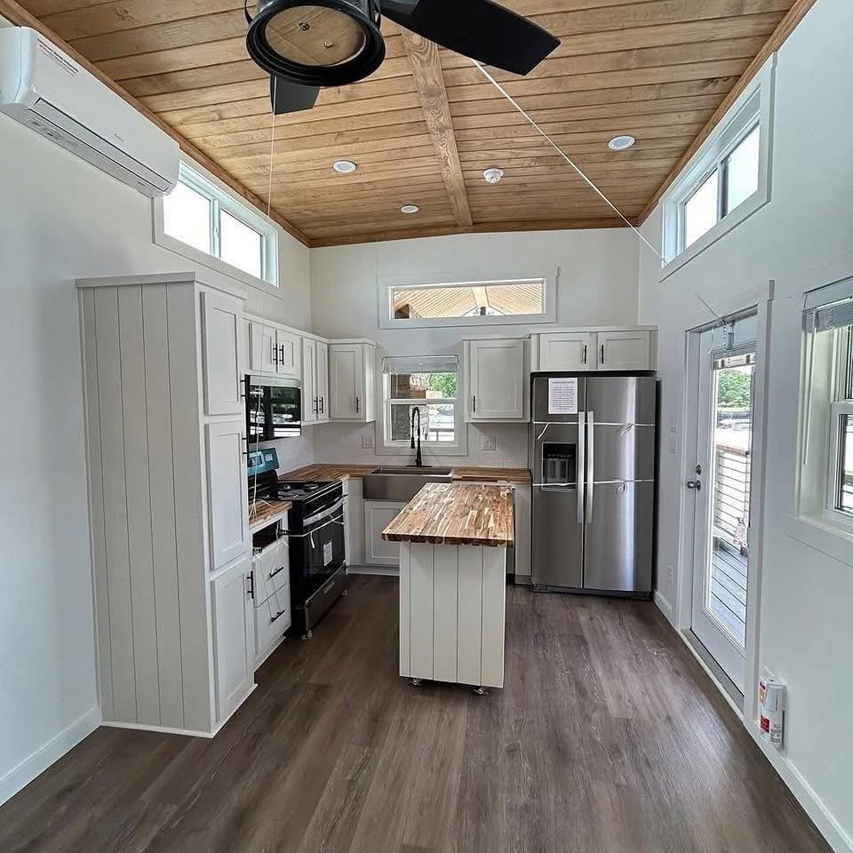 Modern kitchen with white cabinetry, wooden countertops, and stainless steel appliances. It features a wooden ceiling, gray flooring, and large windows, creating a bright and cozy atmosphere.