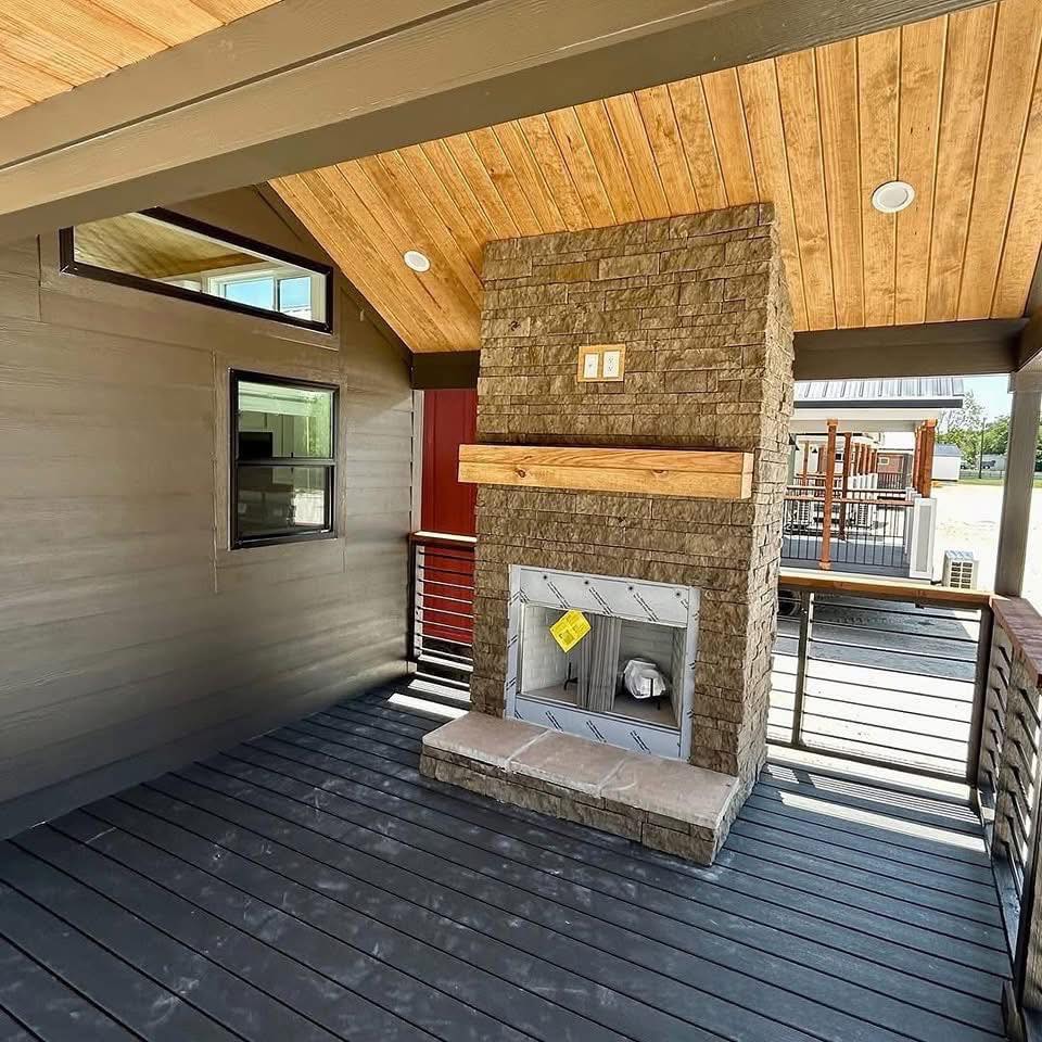 Covered patio with wood-paneled ceiling, featuring a stone fireplace and railing. Soft natural light and warm tones convey a cozy atmosphere.