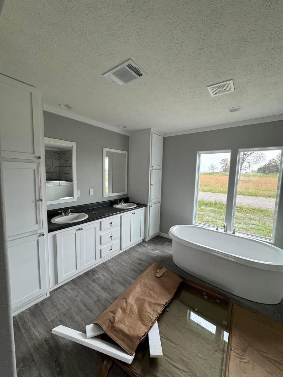 Modern bathroom with a double-sink vanity, sleek black countertop, and white cabinets. A freestanding tub overlooks a yard through large windows.