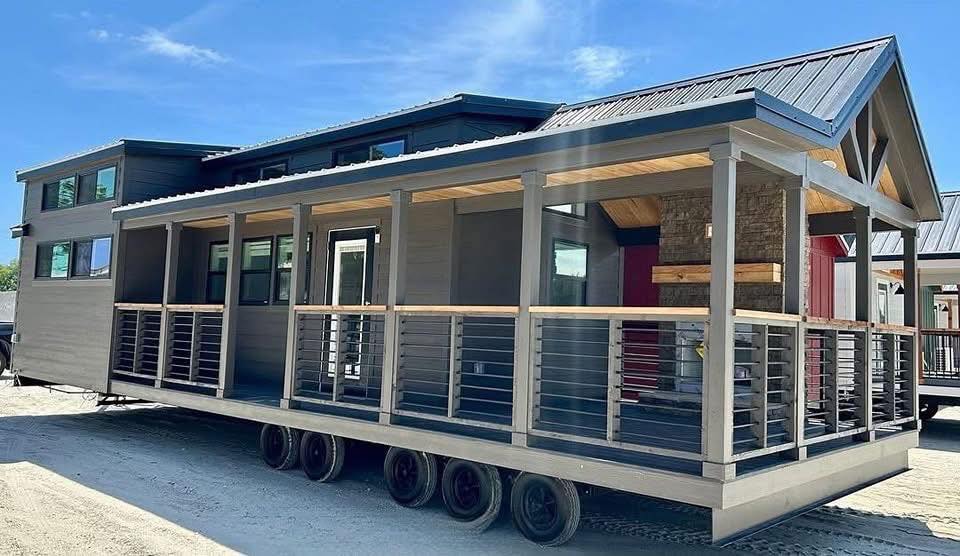 Mobile home with a porch on wheels, featuring modern gray siding and a slanted roof, set under a clear blue sky, conveying a sense of mobility.