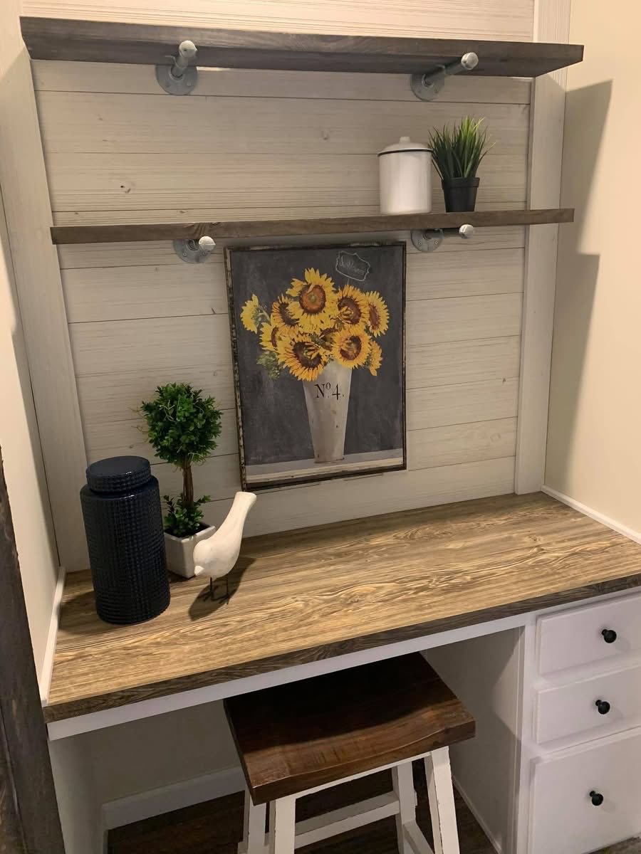 Cozy nook with wooden desk, shelves, and a sunflower painting. Decor includes a small plant, white canister, bird figurine, and dark vase, creating a rustic charm.