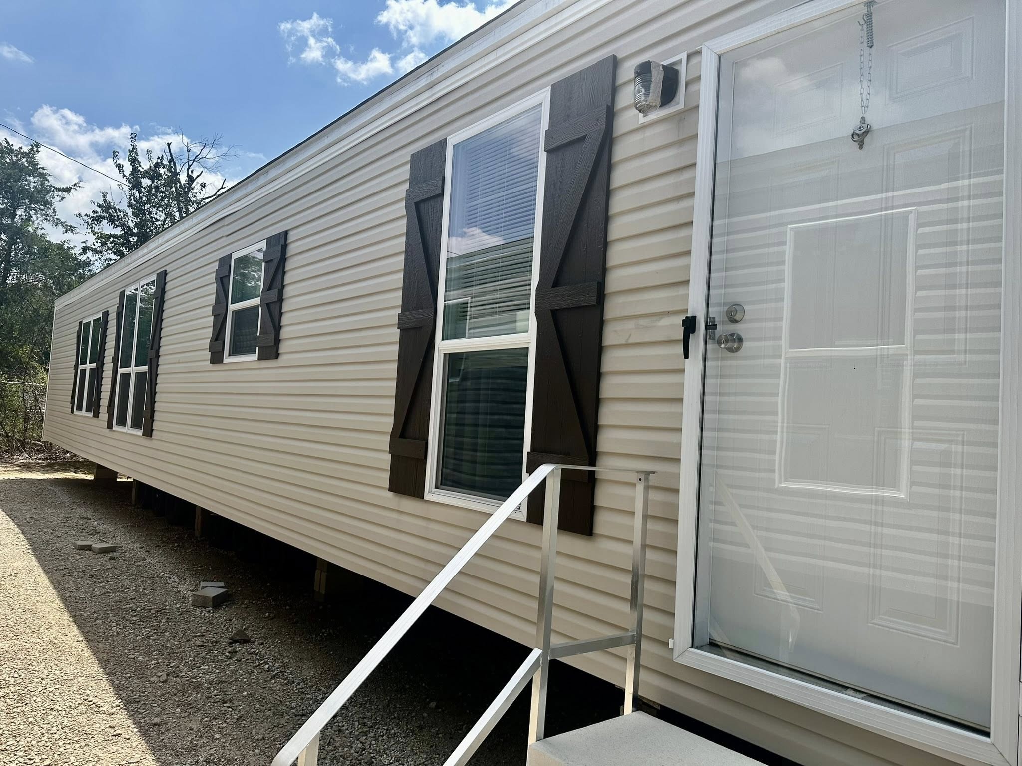 Single-story beige mobile home with brown shutters and multiple windows. A small set of steps leads to the front door. Bright, clear sky above.