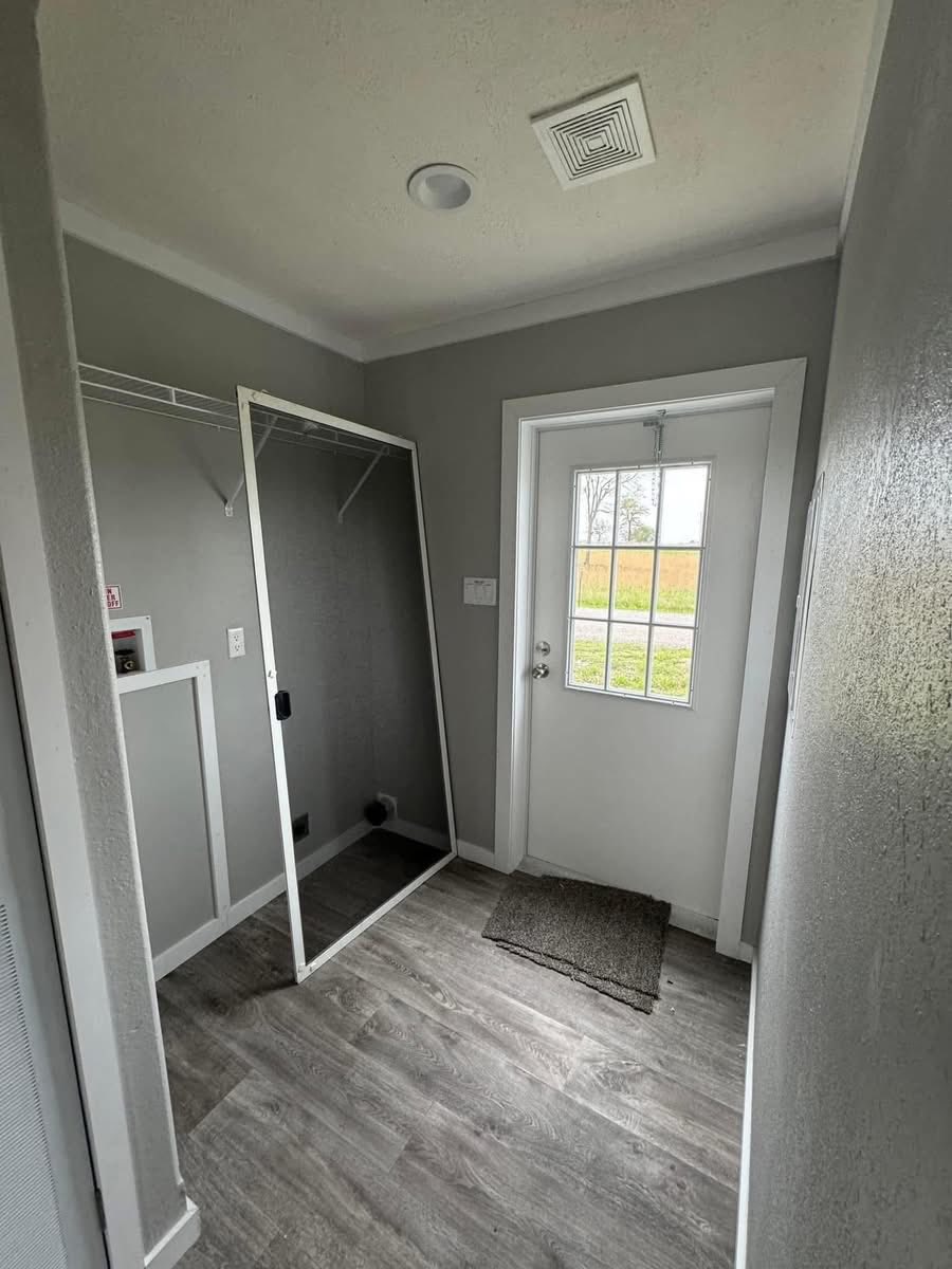 Narrow room with gray walls, wooden floor, a door with window panes, and a leaning mirror. A small mat and closet area add a minimalistic feel.