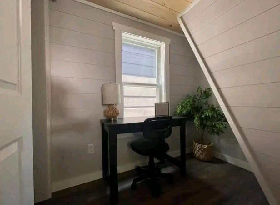 Cozy home office with a black desk and chair, positioned under a window. A lamp and potted plant add warmth. Sloped ceiling creates an intimate feel.