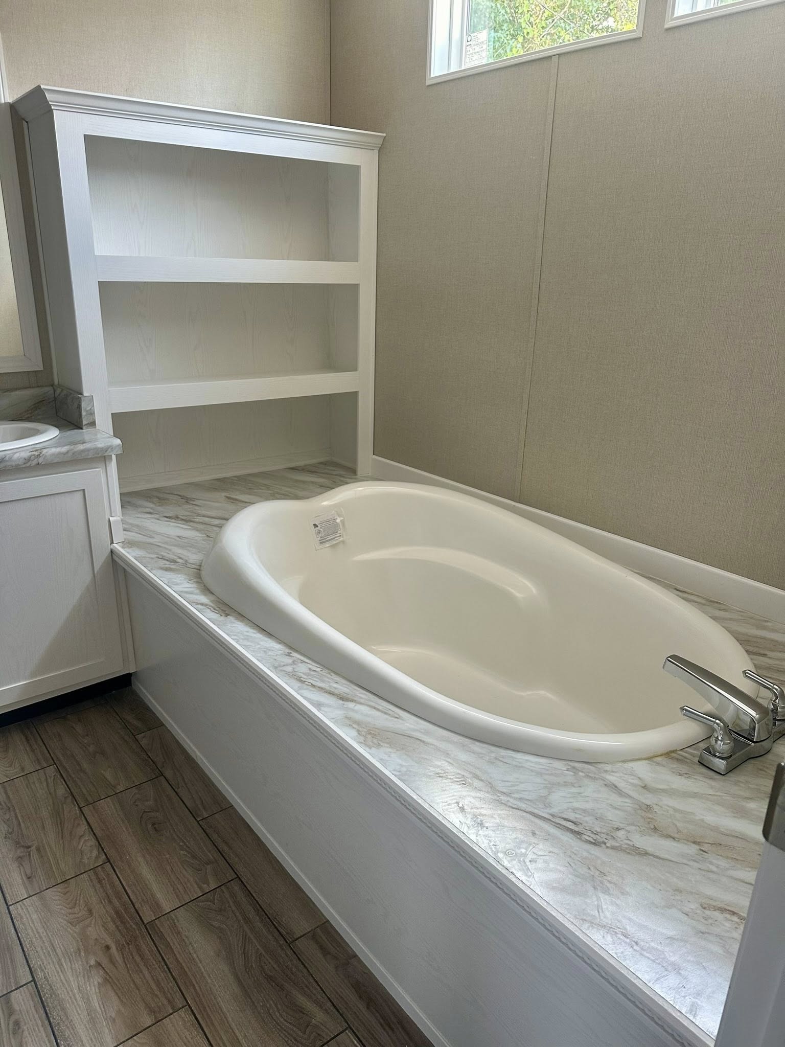 White bathroom with a large, oval bathtub set in a marble-look surround. Wooden shelves and beige walls create a calm, minimalist atmosphere.