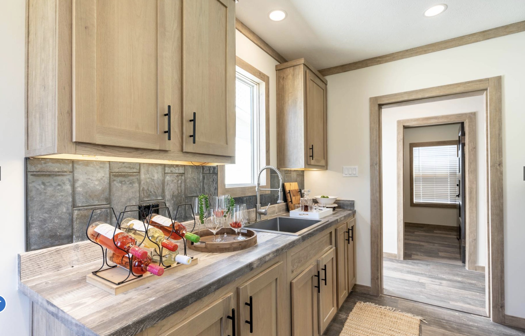A modern kitchen features light wooden cabinets, a sleek countertop with a wine rack and glasses, and a metallic sink. The room is well-lit and inviting.