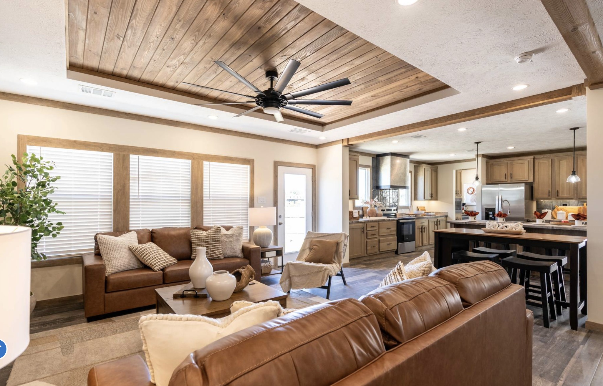 Stylish open-concept living room with brown leather sofas, wood-accented ceiling, and modern kitchen. Natural light and cozy decor create a warm ambiance.