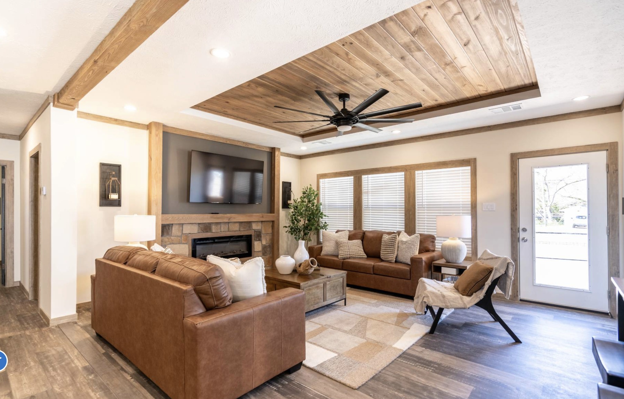 A cozy living room with two brown leather couches, a large TV above a fireplace, wooden ceiling accents, a ceiling fan, and large windows. Warm, inviting atmosphere.