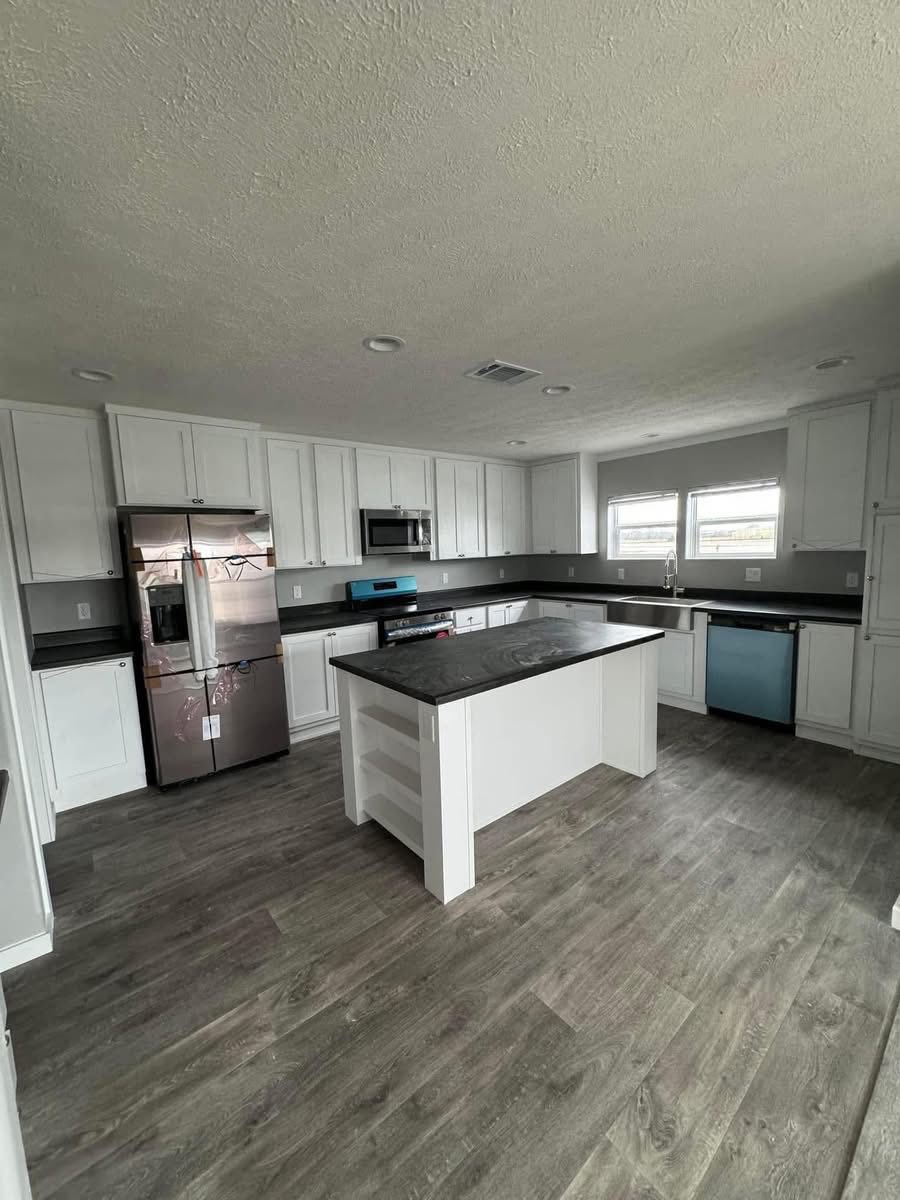 Modern kitchen with white cabinets, dark countertops, and stainless steel appliances. Features a central island and wooden floors under soft lighting.