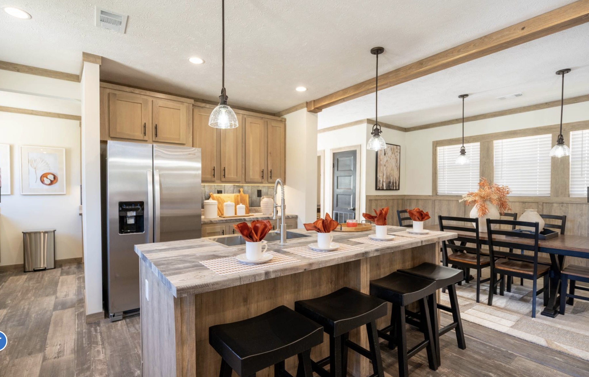 Modern kitchen with wooden cabinetry, island with four black stools, pendant lighting, and a dining area. Bright, warm tones create an inviting feel.