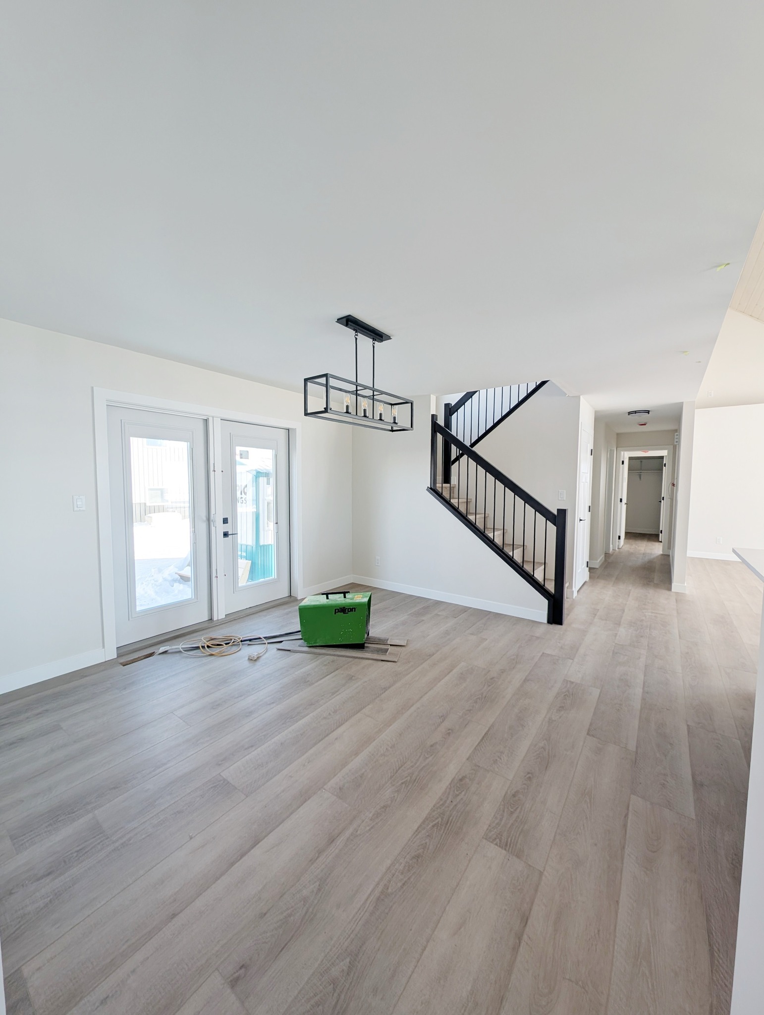 Bright, modern room with light wood floors, white walls, and a black staircase railing. A large black chandelier hangs, and tools lie near glass doors.