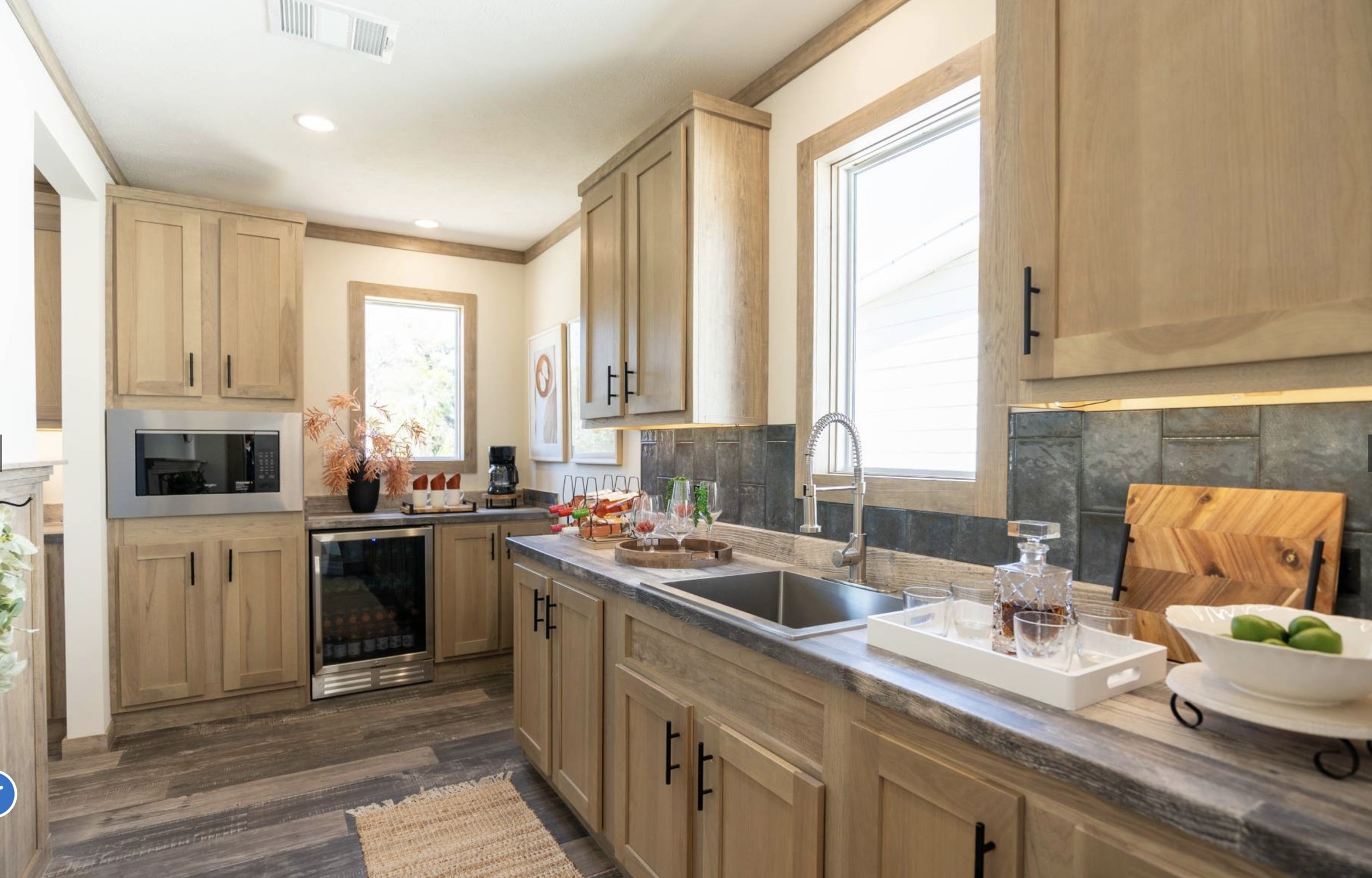 Modern kitchen with light wood cabinets and dark countertops. Sunlight from a window illuminates a clean space with neatly arranged drinks, a wine fridge, and plants, conveying a warm and inviting atmosphere.
