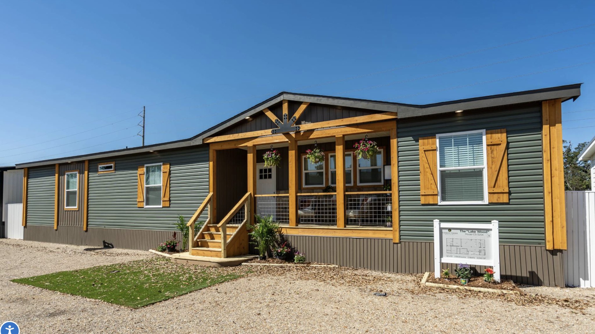 A modern modular home with green siding and wooden trim under a clear blue sky. The porch features potted plants, and a white sign stands nearby.