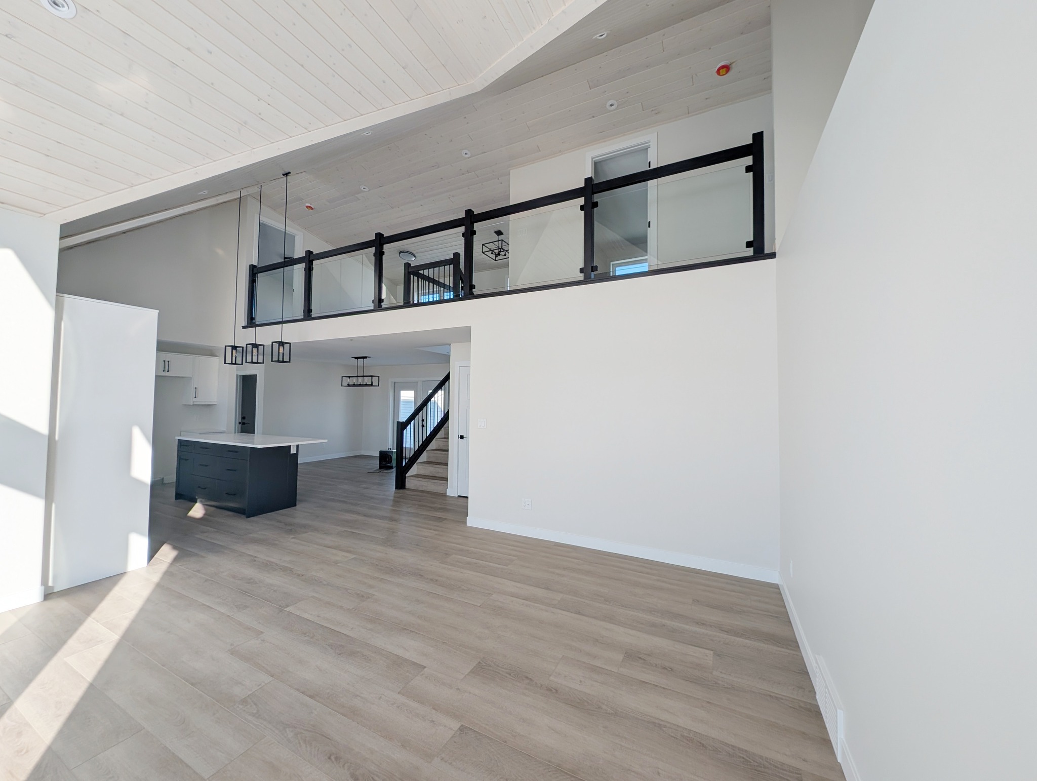 Spacious modern interior with light wood floors, high ceiling, and large windows. Features a sleek kitchen with dark island and an open loft area.