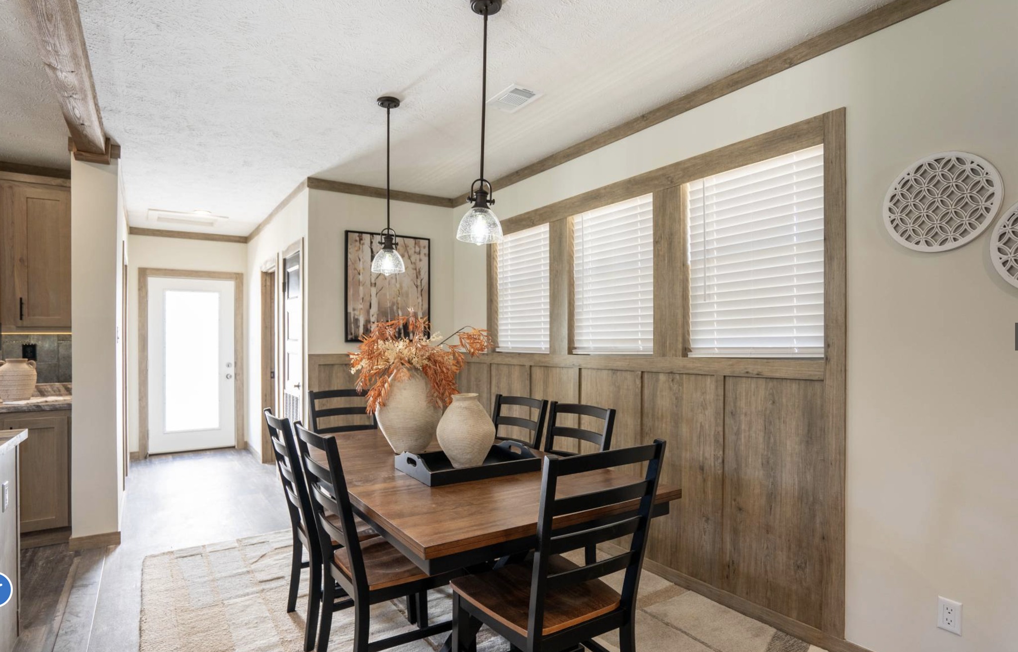 Dining area with a rustic wooden table set for six, two ceramic vases with dried flowers as a centerpiece, pendant lights, and three windows with blinds. Cozy and inviting atmosphere.