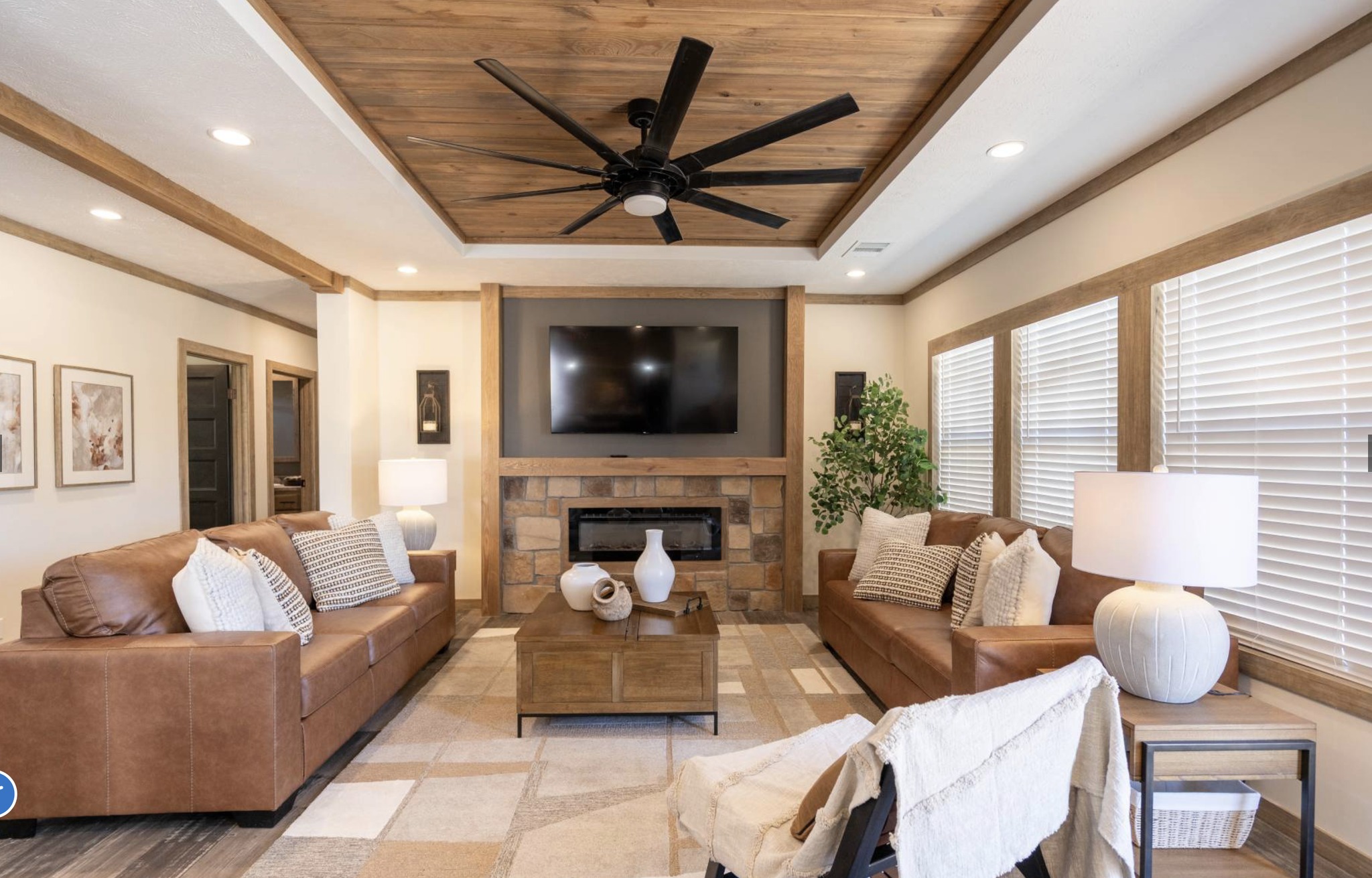 A cozy living room with two brown leather sofas facing each other, a wooden coffee table in between, and a stone fireplace with a TV above. Warm lighting and a ceiling fan enhance the inviting atmosphere.