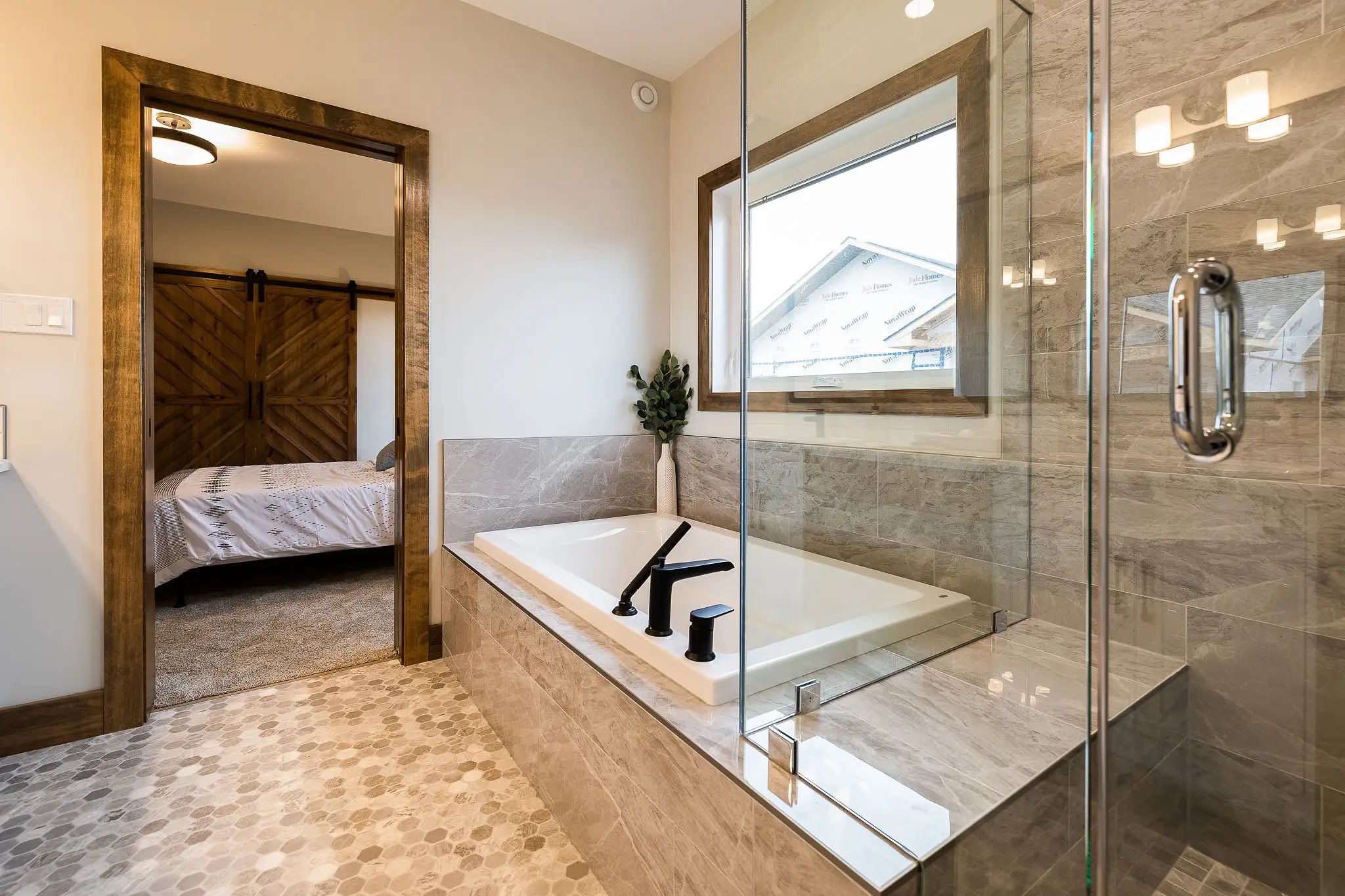 Modern bathroom with glass shower, rectangular tub, and sleek black fixtures. Warm tones, soft lighting, and view into a cozy bedroom with a barn door.