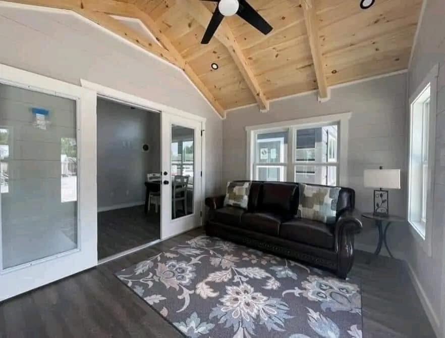 Cozy living room with vaulted wood ceiling, dark leather sofa, floral patterned rug, and large windows. Bright and inviting with modern decor.