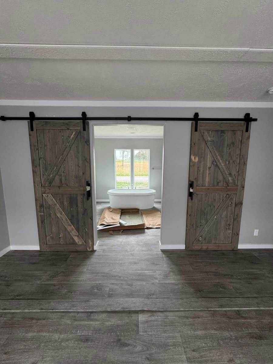 Rustic sliding barn doors open to a bathroom with a freestanding tub by a large window. The room features neutral tones and a serene atmosphere.