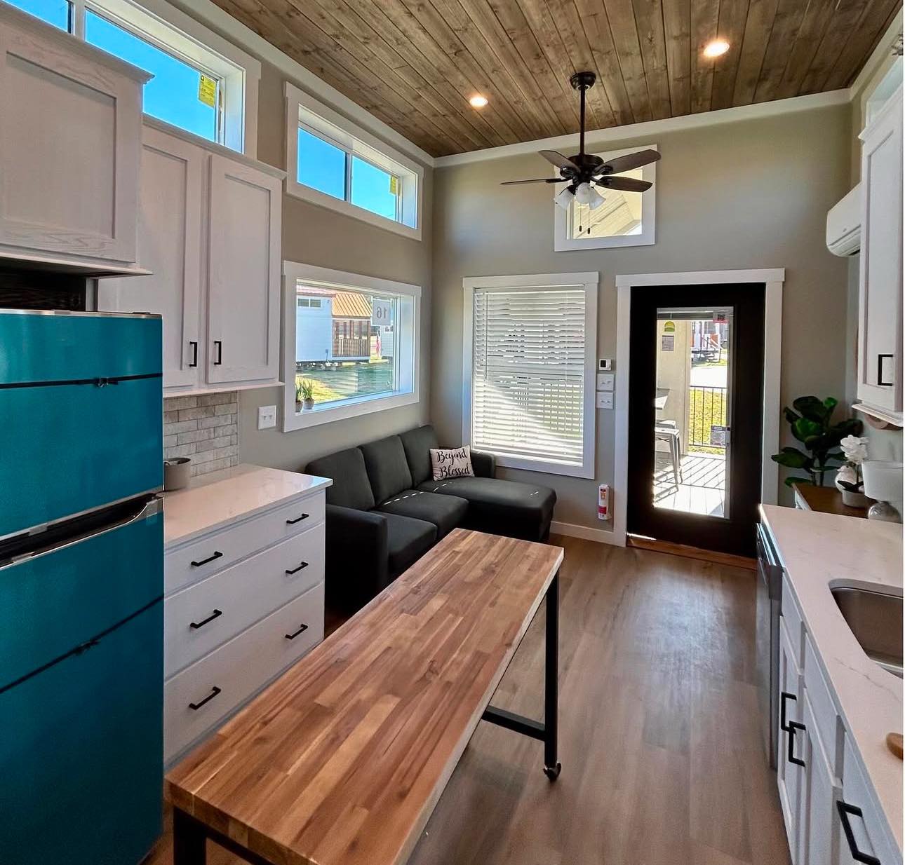 Cozy tiny home interior with a teal fridge, wooden island, and gray sofa. Bright windows, wood ceiling, and door add warmth. Modern and inviting.