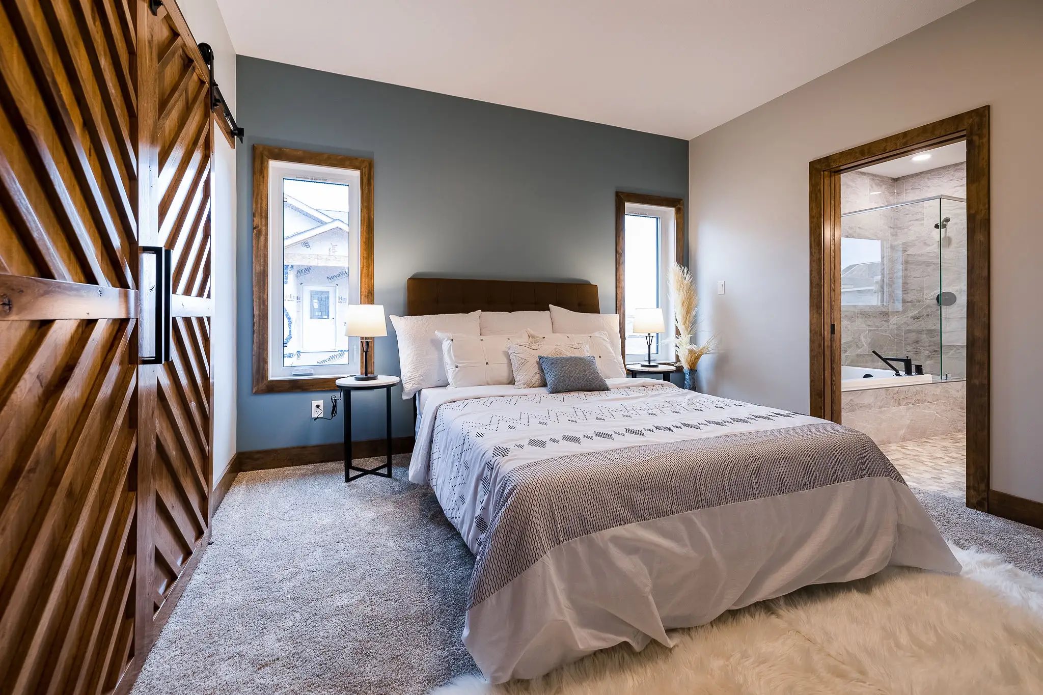 Cozy bedroom with blue accent wall, a soft bed, and plush rug. Wooden sliding door on the left, modern bathroom visible to the right. Warm, inviting ambiance.