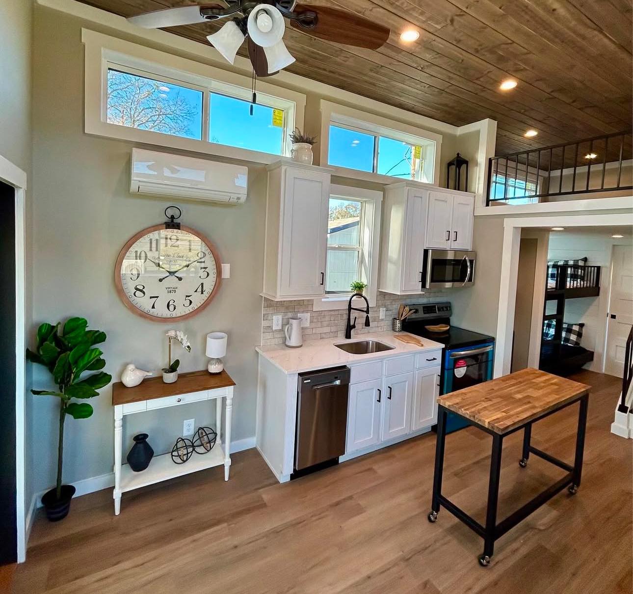 A cozy tiny home kitchen features white cabinets, stainless steel appliances, a wooden island, large wall clock, ceiling fan, and loft with a modern design.