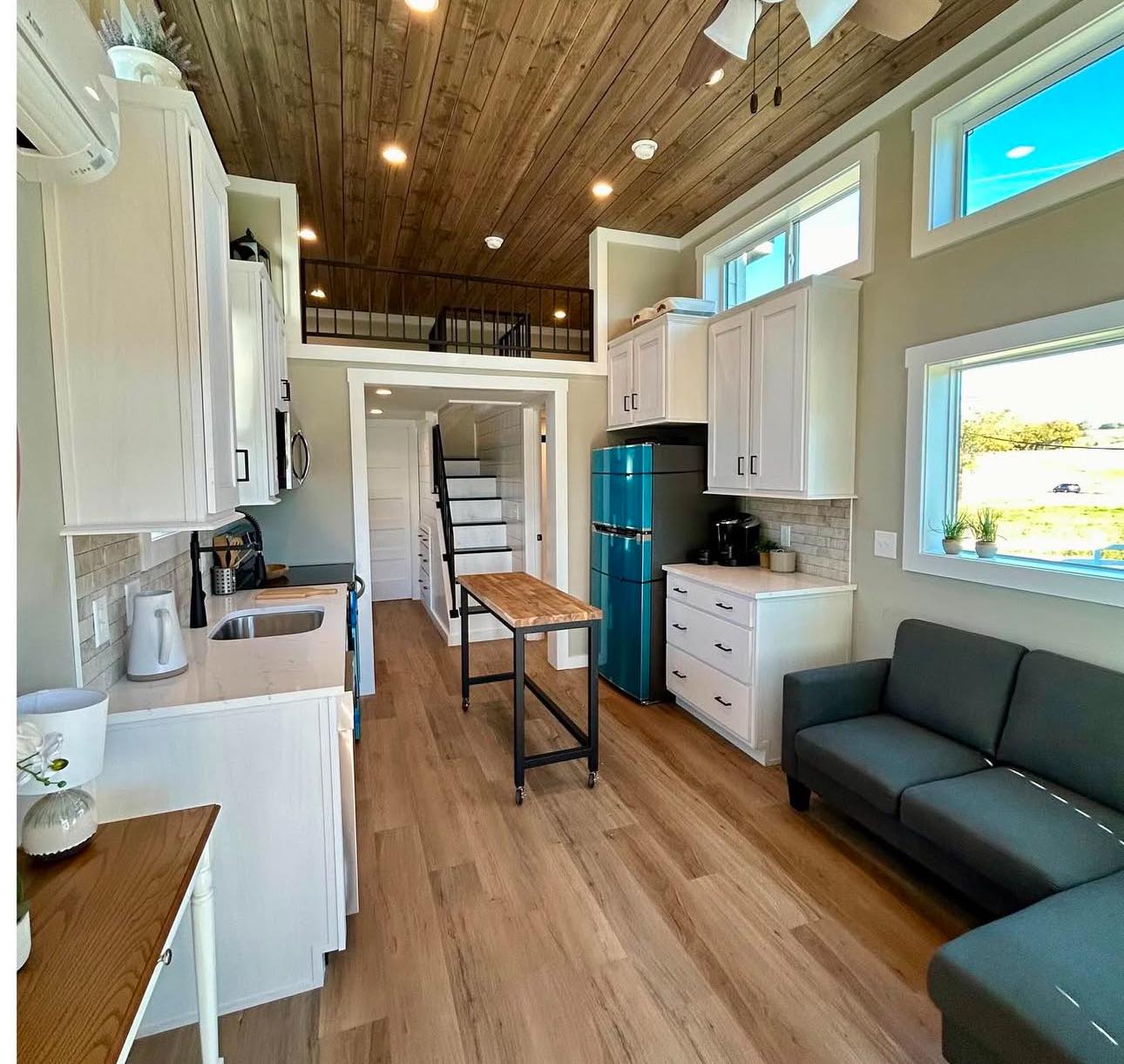 Cozy tiny house interior with wood ceiling and floors. Features a small kitchen, teal fridge, gray sofa, loft area, and natural light from large windows.