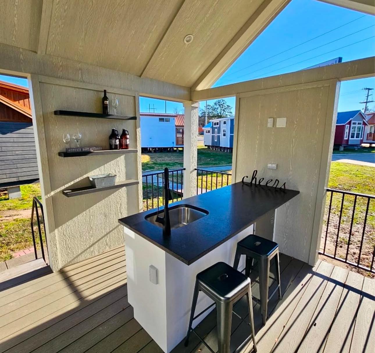 Outdoor kitchen with a sleek black countertop, bar stools, and shelves holding glassware and bottles. Sunlit, cozy, and inviting atmosphere.