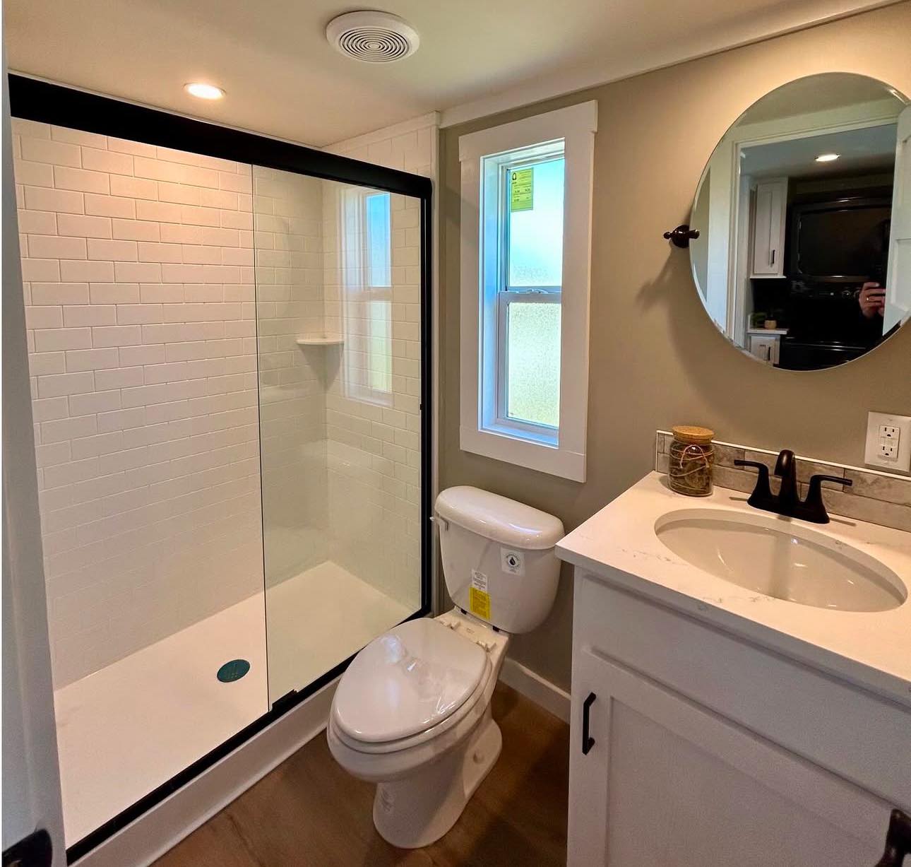 Modern bathroom with a sleek shower featuring white tiles and glass doors, a toilet, and a white vanity with a round mirror above. Bright and clean.