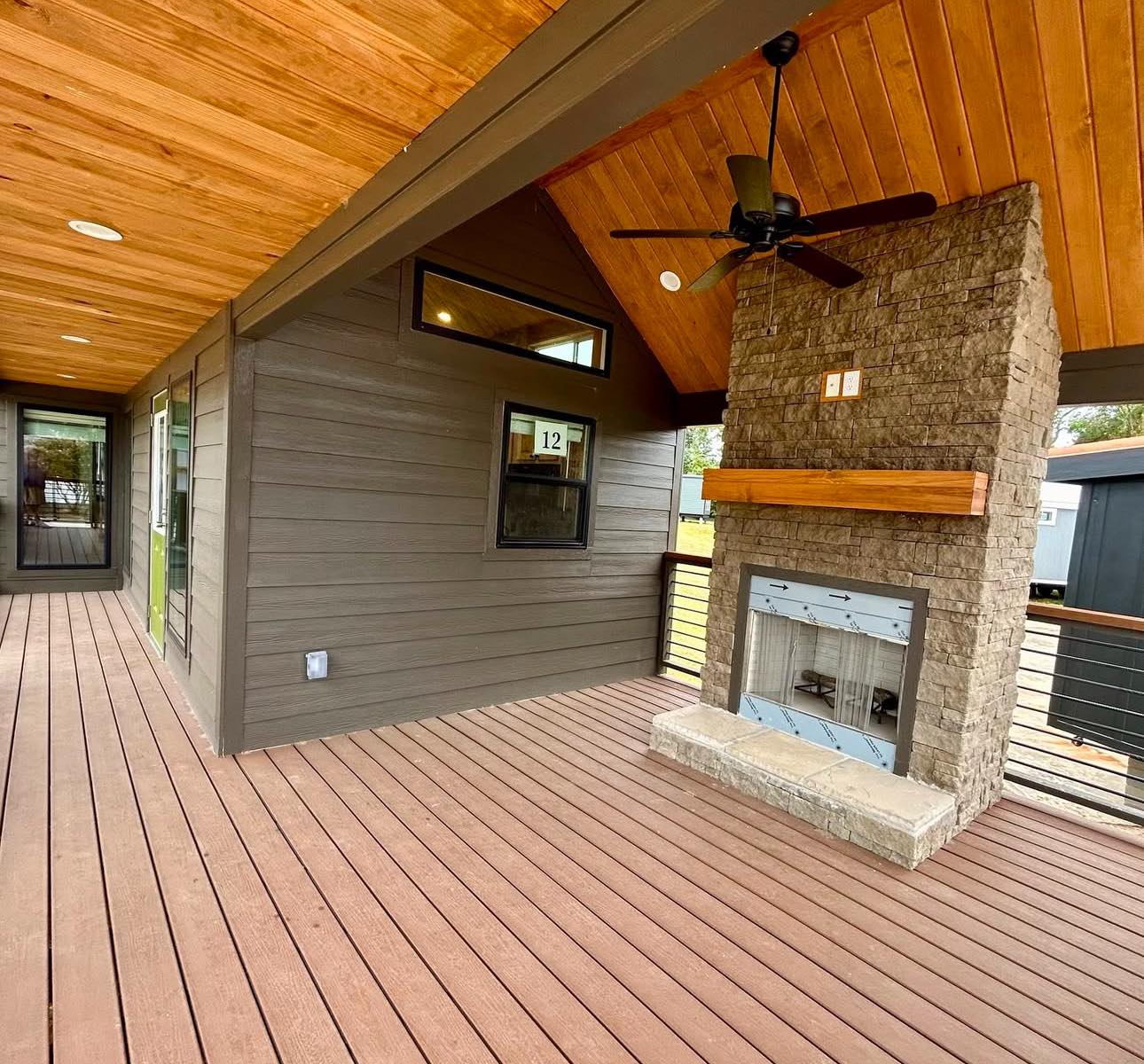 Covered patio with wooden decking and ceiling, featuring a stone fireplace and ceiling fan. Modern, cozy ambiance with natural light.