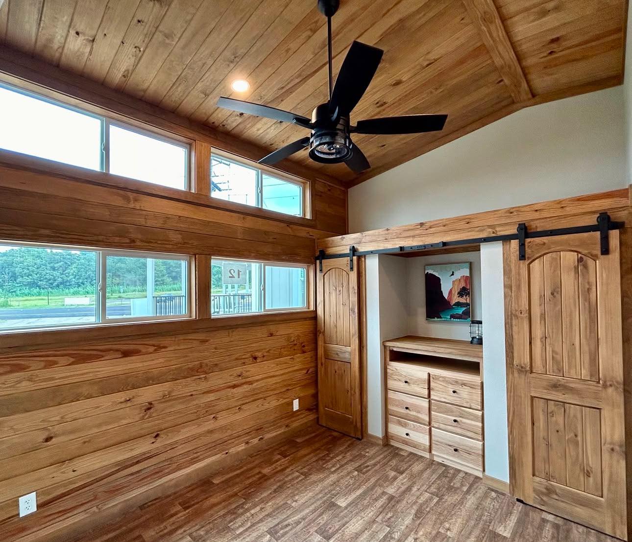 Wood-paneled room with vaulted ceiling, skylights, and a black ceiling fan. Features sliding barn doors, a built-in dresser, and a landscape painting. Cozy and rustic.