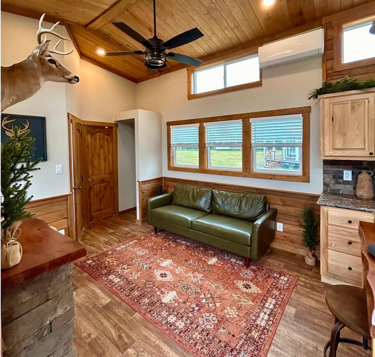 Cozy rustic living room with wood-paneled walls and ceiling, featuring a green leather sofa, red patterned rug, deer mount, and modern ceiling fan.