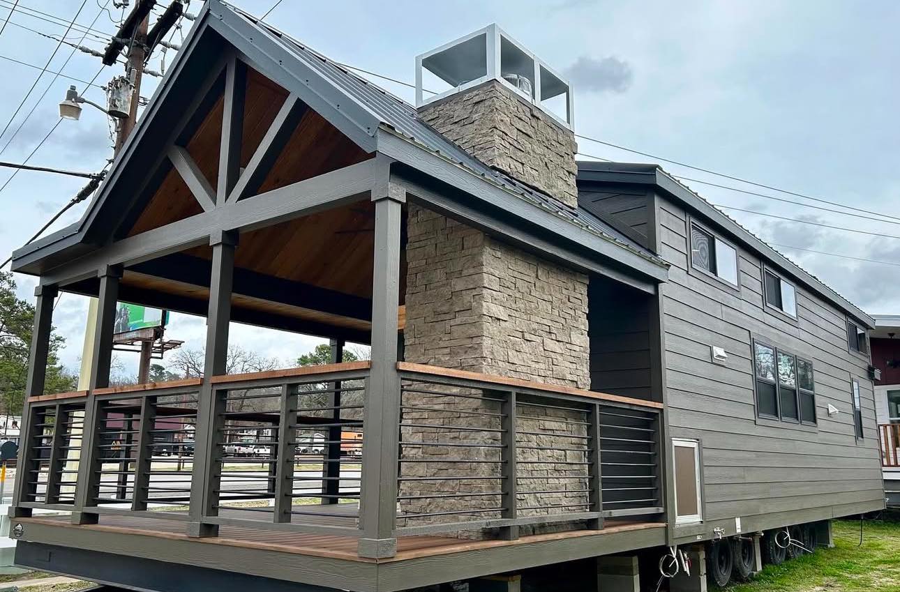 Modern tiny house on wheels with gray siding, large stone chimney, and spacious porch. Overcast sky adds a cozy, inviting feel to the scene.
