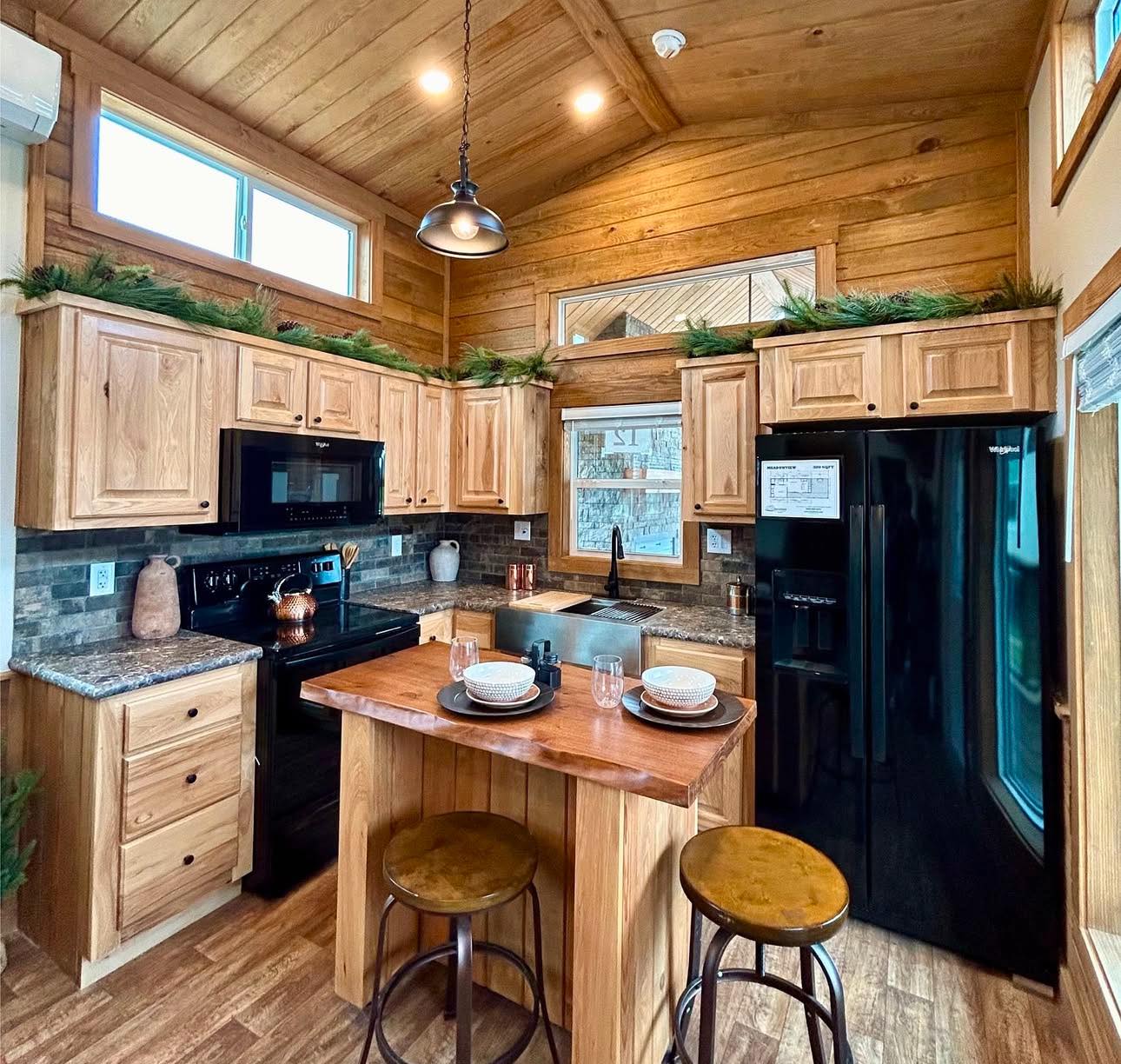 Cozy kitchen with wooden cabinets, black appliances, and a central island with stools. Greenery decorates the tops, adding a rustic touch. Warm lighting creates an inviting atmosphere.