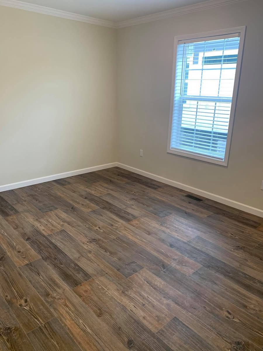 Empty room with light beige walls and dark wood flooring. A single window with white blinds allows natural light, creating a calm and inviting space.